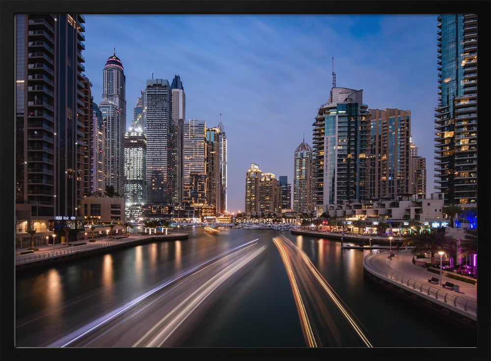 A long-exposure photograph of the Dubai Marina at dusk, framed in silver. Illuminated skyscrapers line a busy waterway, with light trails from moving boats creating long, bright streaks on the water. The sky is a deep twilight blue, and the city's lights cast a warm glow. Decor