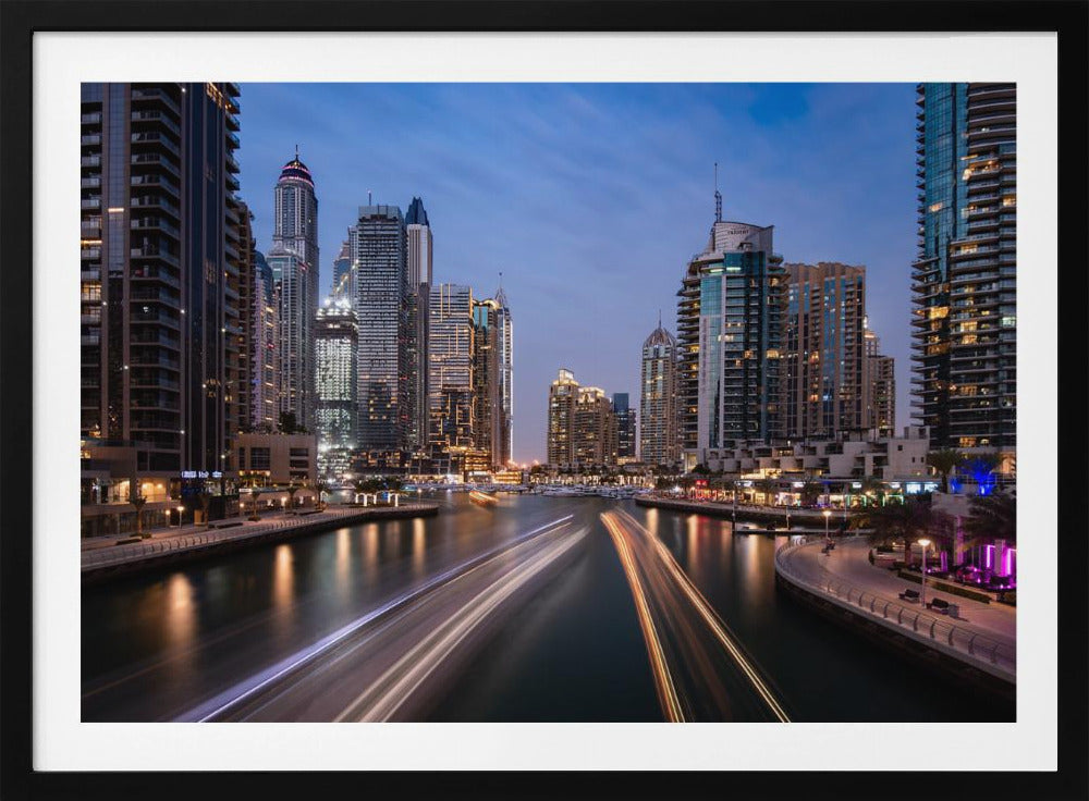 A long-exposure photograph of the Dubai Marina at dusk, framed in silver. Illuminated skyscrapers line a busy waterway, with light trails from moving boats creating long, bright streaks on the water. The sky is a deep twilight blue, and the city's lights cast a warm glow. Decor