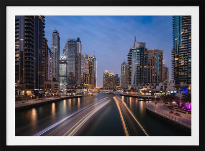 A long-exposure photograph of the Dubai Marina at dusk, framed in silver. Illuminated skyscrapers line a busy waterway, with light trails from moving boats creating long, bright streaks on the water. The sky is a deep twilight blue, and the city's lights cast a warm glow. Decor
