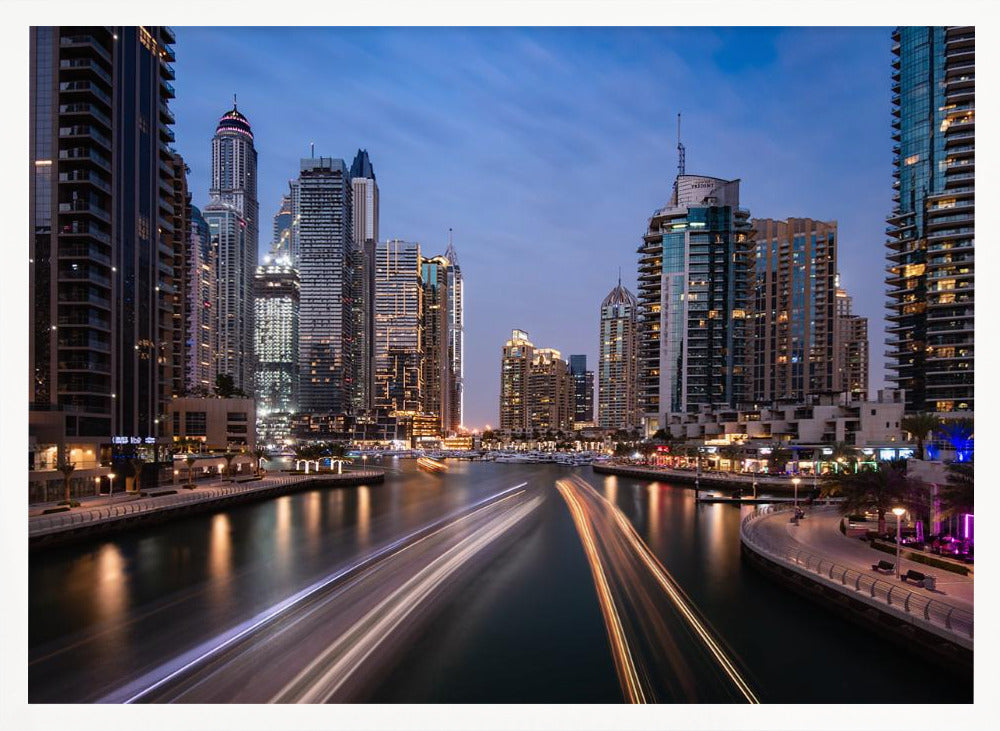 A long-exposure photograph of the Dubai Marina at dusk, framed in silver. Illuminated skyscrapers line a busy waterway, with light trails from moving boats creating long, bright streaks on the water. The sky is a deep twilight blue, and the city's lights cast a warm glow. Decor