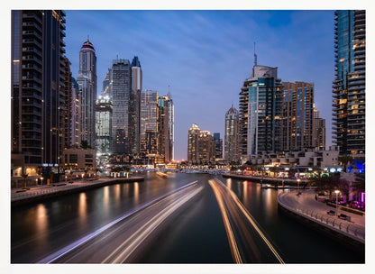 A long-exposure photograph of the Dubai Marina at dusk, framed in silver. Illuminated skyscrapers line a busy waterway, with light trails from moving boats creating long, bright streaks on the water. The sky is a deep twilight blue, and the city's lights cast a warm glow. Decor
