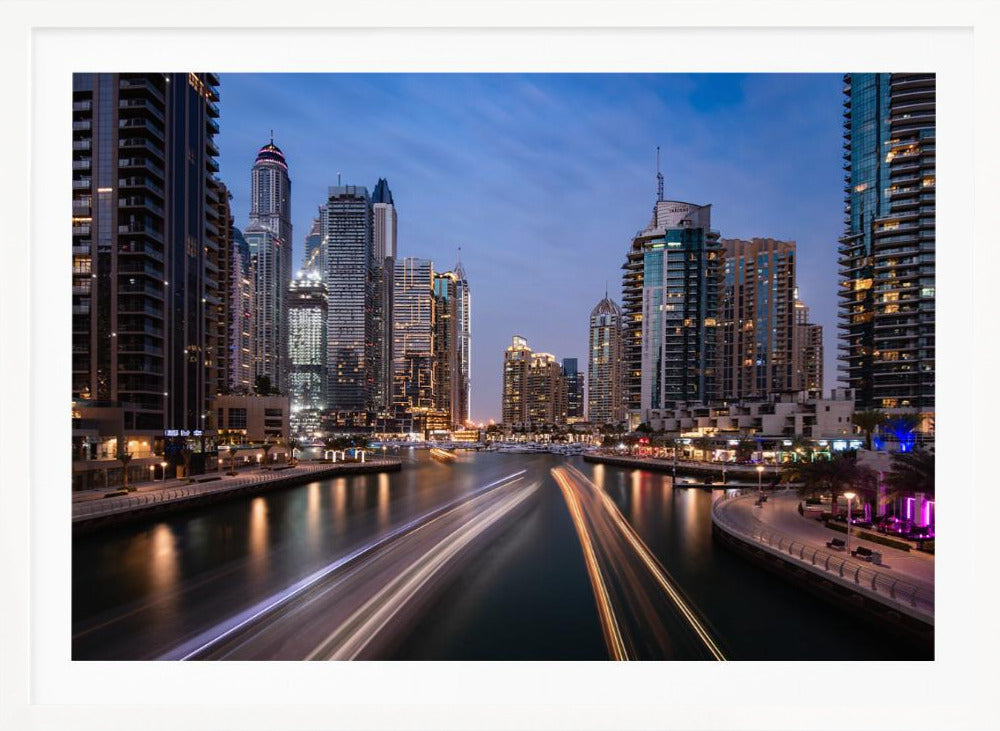 A long-exposure photograph of the Dubai Marina at dusk, framed in silver. Illuminated skyscrapers line a busy waterway, with light trails from moving boats creating long, bright streaks on the water. The sky is a deep twilight blue, and the city's lights cast a warm glow. Decor