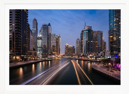 A long-exposure photograph of the Dubai Marina at dusk, framed in silver. Illuminated skyscrapers line a busy waterway, with light trails from moving boats creating long, bright streaks on the water. The sky is a deep twilight blue, and the city's lights cast a warm glow. Decor
