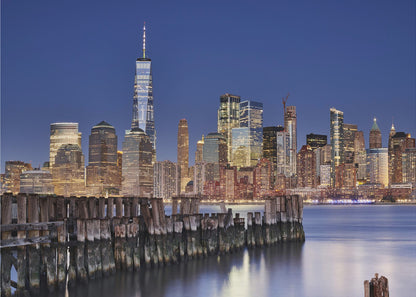 A framed photograph of the Lower Manhattan skyline at dusk. The One World Trade Center and other skyscrapers are brightly lit against a deep blue sky. In the foreground, an old wooden pier extends into the water, which reflects the city lights. Wall Art