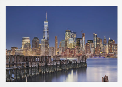 A framed photograph of the Lower Manhattan skyline at dusk. The One World Trade Center and other skyscrapers are brightly lit against a deep blue sky. In the foreground, an old wooden pier extends into the water, which reflects the city lights. Wall Art