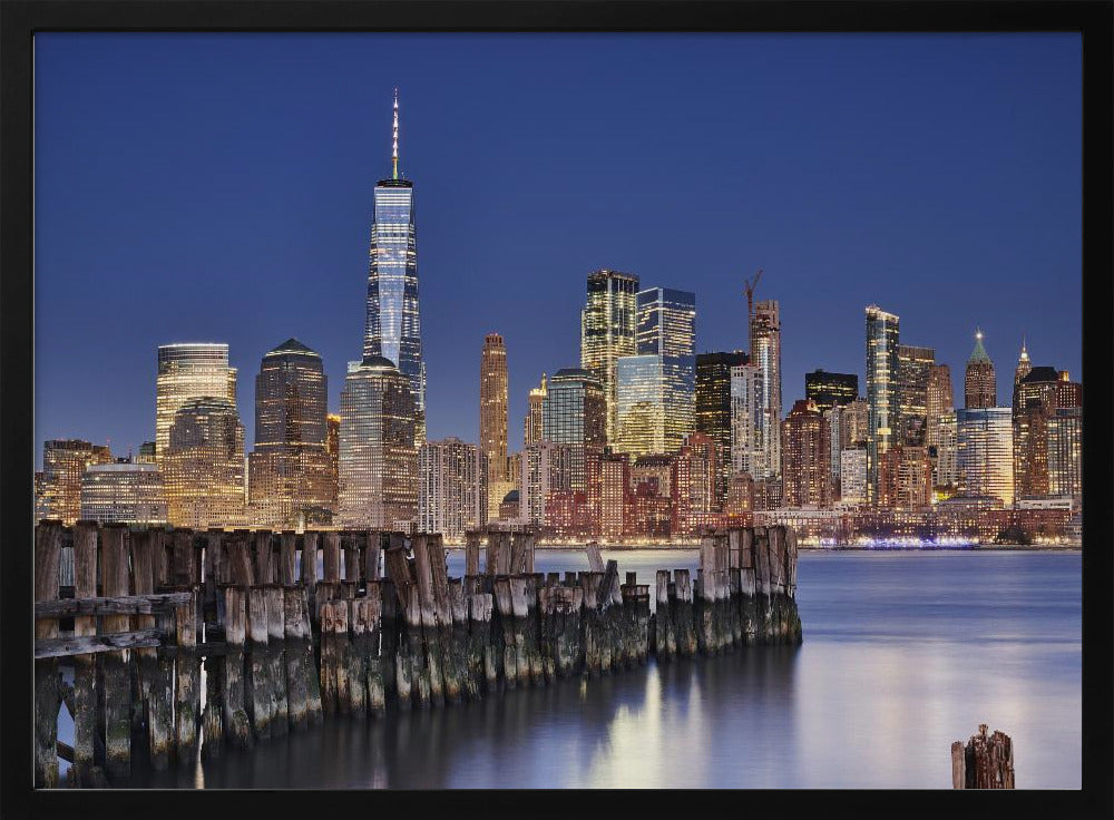 A framed photograph of the Lower Manhattan skyline at dusk. The One World Trade Center and other skyscrapers are brightly lit against a deep blue sky. In the foreground, an old wooden pier extends into the water, which reflects the city lights. Wall Art