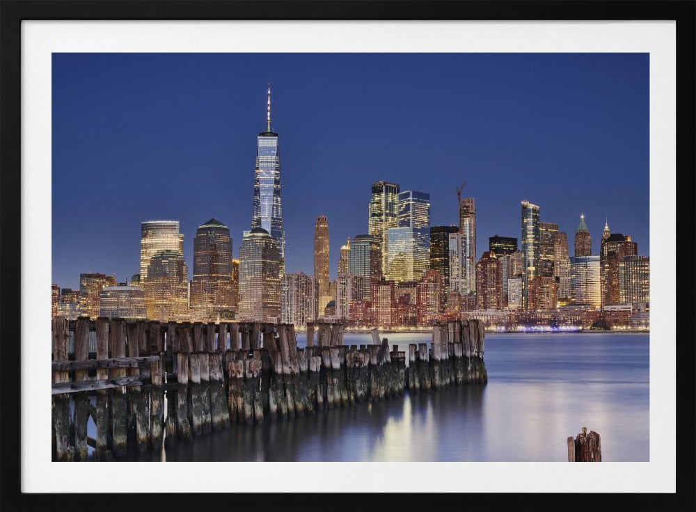 A framed photograph of the Lower Manhattan skyline at dusk. The One World Trade Center and other skyscrapers are brightly lit against a deep blue sky. In the foreground, an old wooden pier extends into the water, which reflects the city lights. Wall Art
