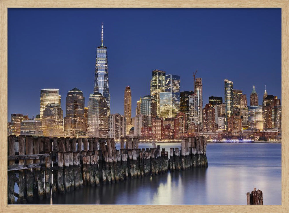 A framed photograph of the Lower Manhattan skyline at dusk. The One World Trade Center and other skyscrapers are brightly lit against a deep blue sky. In the foreground, an old wooden pier extends into the water, which reflects the city lights. Wall Art