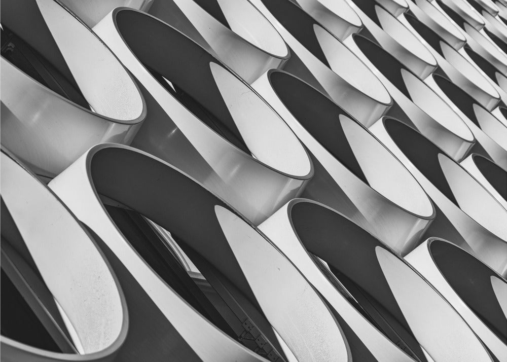 A framed, black and white photograph captures a low-angle, close-up view of a modern building's facade. The image features a repeating pattern of slanted, oval, or teardrop-shaped metallic elements, creating a dynamic interplay of light, shadow, and reflection. Print