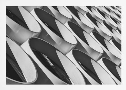 A framed, black and white photograph captures a low-angle, close-up view of a modern building's facade. The image features a repeating pattern of slanted, oval, or teardrop-shaped metallic elements, creating a dynamic interplay of light, shadow, and reflection. Print