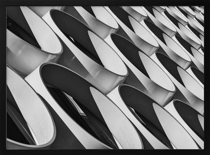 A framed, black and white photograph captures a low-angle, close-up view of a modern building's facade. The image features a repeating pattern of slanted, oval, or teardrop-shaped metallic elements, creating a dynamic interplay of light, shadow, and reflection. Print