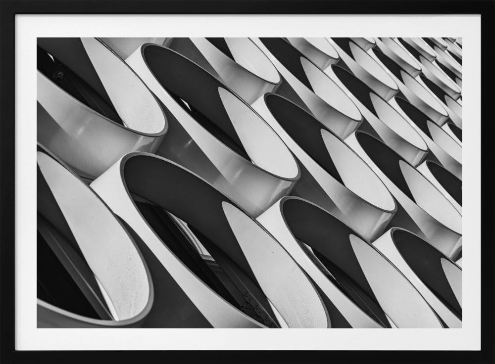 A framed, black and white photograph captures a low-angle, close-up view of a modern building's facade. The image features a repeating pattern of slanted, oval, or teardrop-shaped metallic elements, creating a dynamic interplay of light, shadow, and reflection. Print