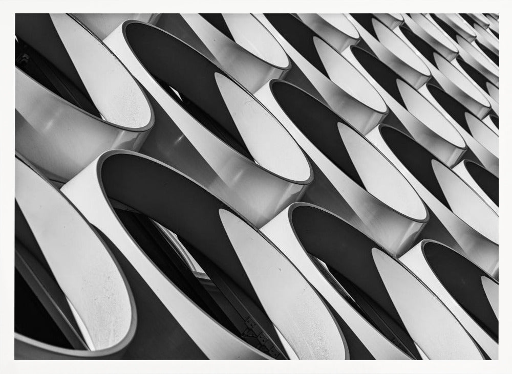 A framed, black and white photograph captures a low-angle, close-up view of a modern building's facade. The image features a repeating pattern of slanted, oval, or teardrop-shaped metallic elements, creating a dynamic interplay of light, shadow, and reflection. Print