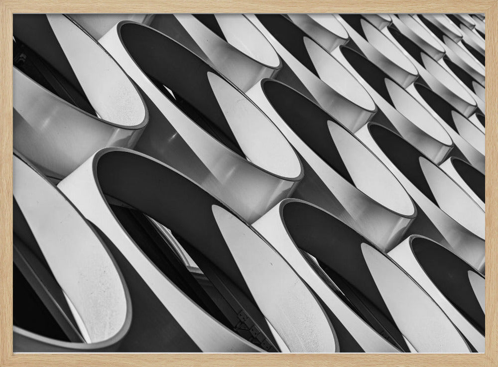 A framed, black and white photograph captures a low-angle, close-up view of a modern building's facade. The image features a repeating pattern of slanted, oval, or teardrop-shaped metallic elements, creating a dynamic interplay of light, shadow, and reflection. Print