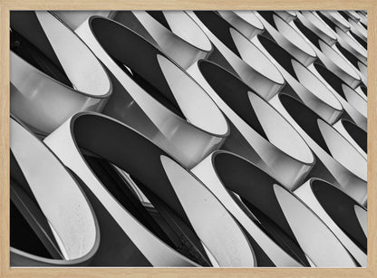 A framed, black and white photograph captures a low-angle, close-up view of a modern building's facade. The image features a repeating pattern of slanted, oval, or teardrop-shaped metallic elements, creating a dynamic interplay of light, shadow, and reflection. Print