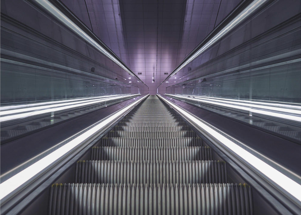 A symmetrical, first-person view looking down a long, empty escalator in a modern subway station, with bright white lights lining the handrails and a purple glow on the ceiling, creating a futuristic and infinite perspective. Wall Art