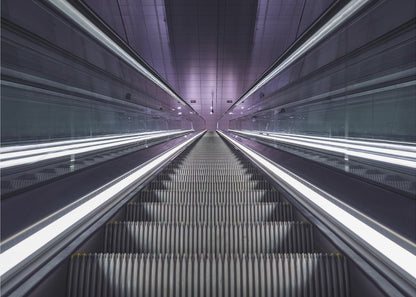 A symmetrical, first-person view looking down a long, empty escalator in a modern subway station, with bright white lights lining the handrails and a purple glow on the ceiling, creating a futuristic and infinite perspective. Wall Art