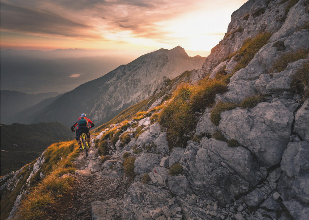 A mountain biker from behind rides along a narrow, rocky trail on a steep mountain ridge during a dramatic golden sunset. In the background, layers of mountains fade into the distance under a warm, glowing sky. Artwork