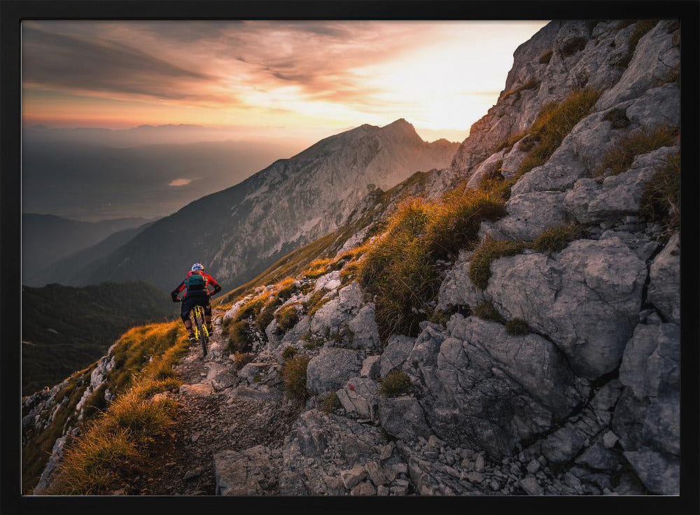 A mountain biker from behind rides along a narrow, rocky trail on a steep mountain ridge during a dramatic golden sunset. In the background, layers of mountains fade into the distance under a warm, glowing sky. Artwork