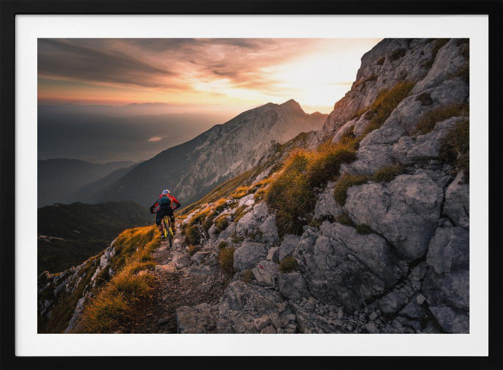 A mountain biker from behind rides along a narrow, rocky trail on a steep mountain ridge during a dramatic golden sunset. In the background, layers of mountains fade into the distance under a warm, glowing sky. Artwork