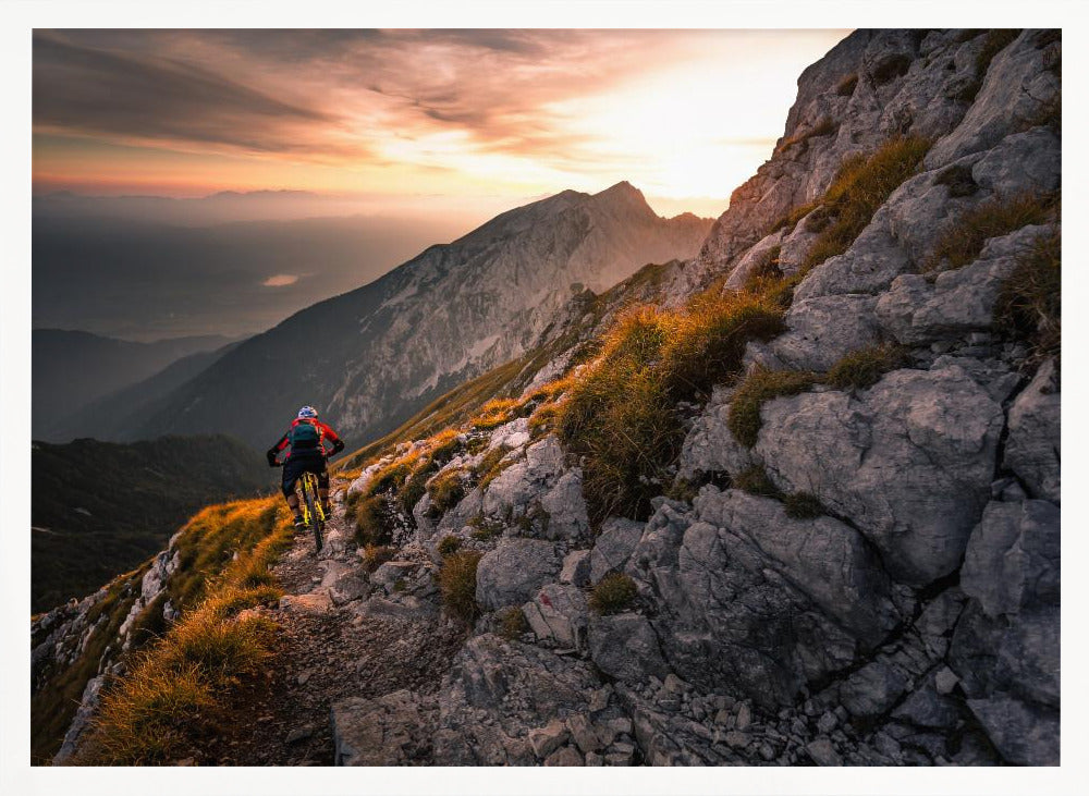 A mountain biker from behind rides along a narrow, rocky trail on a steep mountain ridge during a dramatic golden sunset. In the background, layers of mountains fade into the distance under a warm, glowing sky. Artwork