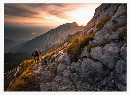 A mountain biker from behind rides along a narrow, rocky trail on a steep mountain ridge during a dramatic golden sunset. In the background, layers of mountains fade into the distance under a warm, glowing sky. Artwork