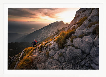 A mountain biker from behind rides along a narrow, rocky trail on a steep mountain ridge during a dramatic golden sunset. In the background, layers of mountains fade into the distance under a warm, glowing sky. Artwork