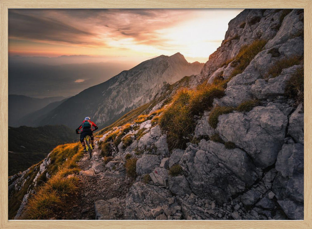 A mountain biker from behind rides along a narrow, rocky trail on a steep mountain ridge during a dramatic golden sunset. In the background, layers of mountains fade into the distance under a warm, glowing sky. Artwork