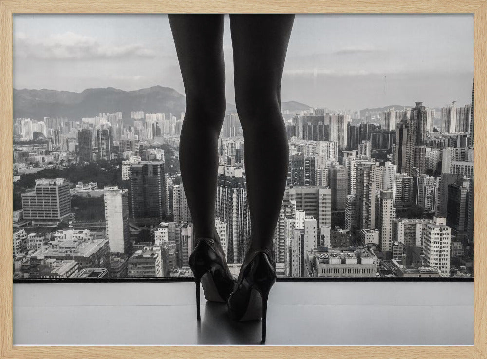 A framed black and white photograph showing the back of a woman's legs and high heels as she stands before a large window, looking out over a vast, dense cityscape with distant mountains under a cloudy sky. Artwork