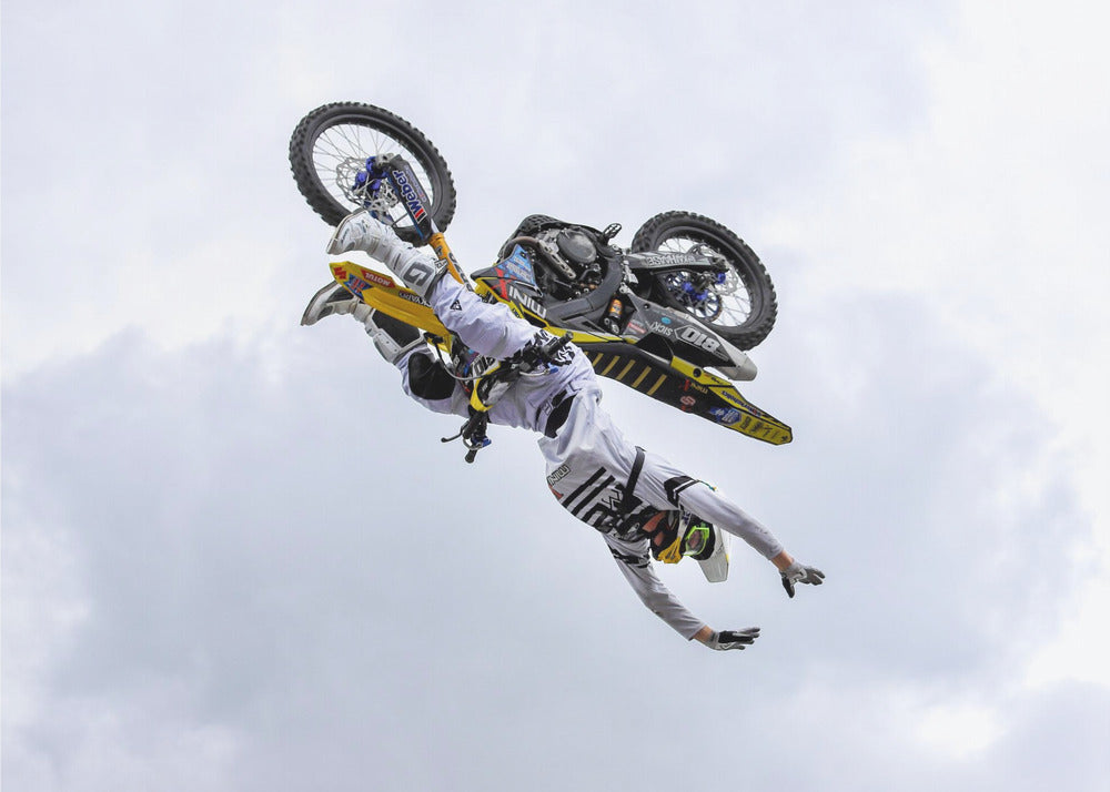 A framed action shot of a freestyle motocross rider in mid-air, performing an upside-down trick against a cloudy sky, with their body hanging below the yellow and black dirt bike. Artwork