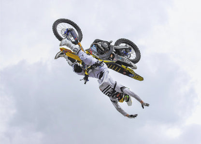A framed action shot of a freestyle motocross rider in mid-air, performing an upside-down trick against a cloudy sky, with their body hanging below the yellow and black dirt bike. Artwork