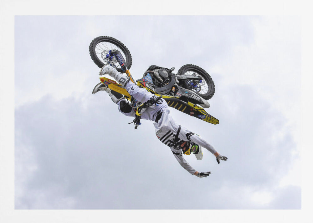 A framed action shot of a freestyle motocross rider in mid-air, performing an upside-down trick against a cloudy sky, with their body hanging below the yellow and black dirt bike. Artwork