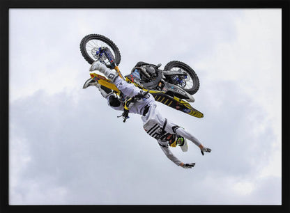 A framed action shot of a freestyle motocross rider in mid-air, performing an upside-down trick against a cloudy sky, with their body hanging below the yellow and black dirt bike. Artwork