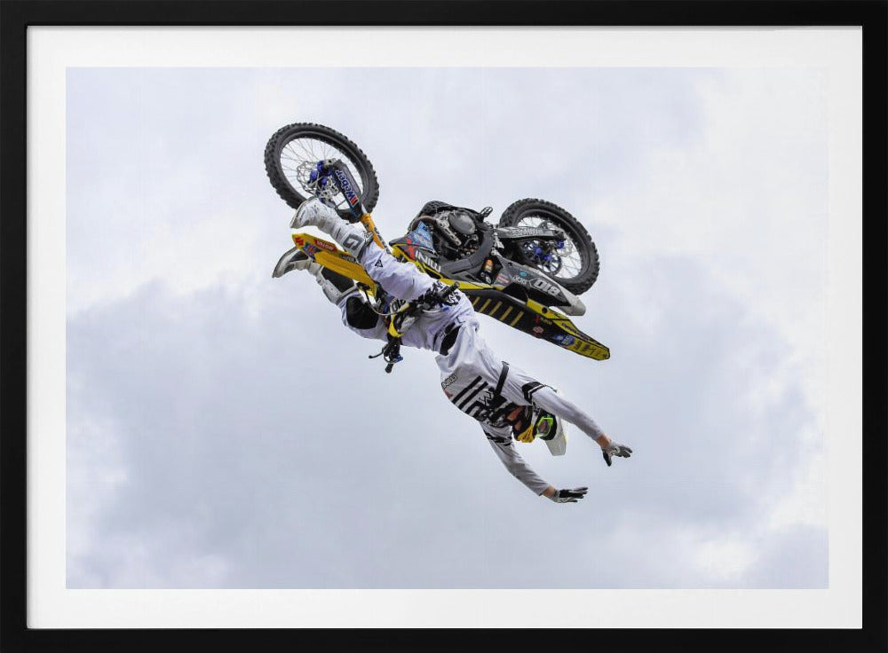 A framed action shot of a freestyle motocross rider in mid-air, performing an upside-down trick against a cloudy sky, with their body hanging below the yellow and black dirt bike. Artwork