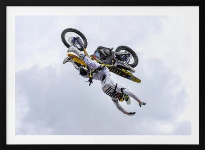A framed action shot of a freestyle motocross rider in mid-air, performing an upside-down trick against a cloudy sky, with their body hanging below the yellow and black dirt bike. Artwork