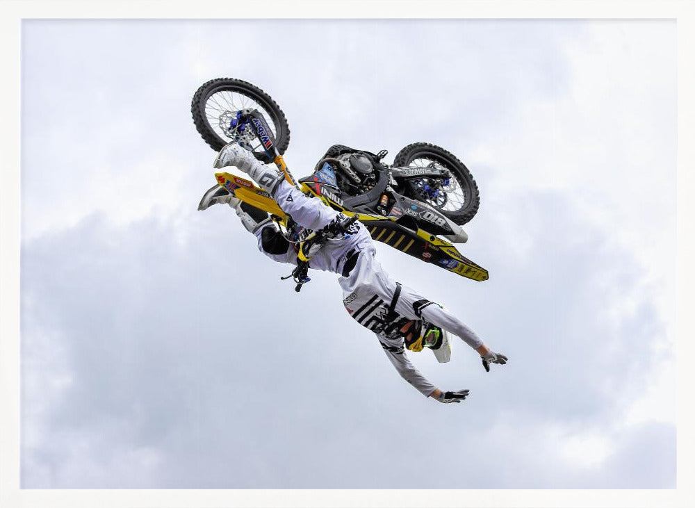 A framed action shot of a freestyle motocross rider in mid-air, performing an upside-down trick against a cloudy sky, with their body hanging below the yellow and black dirt bike. Artwork