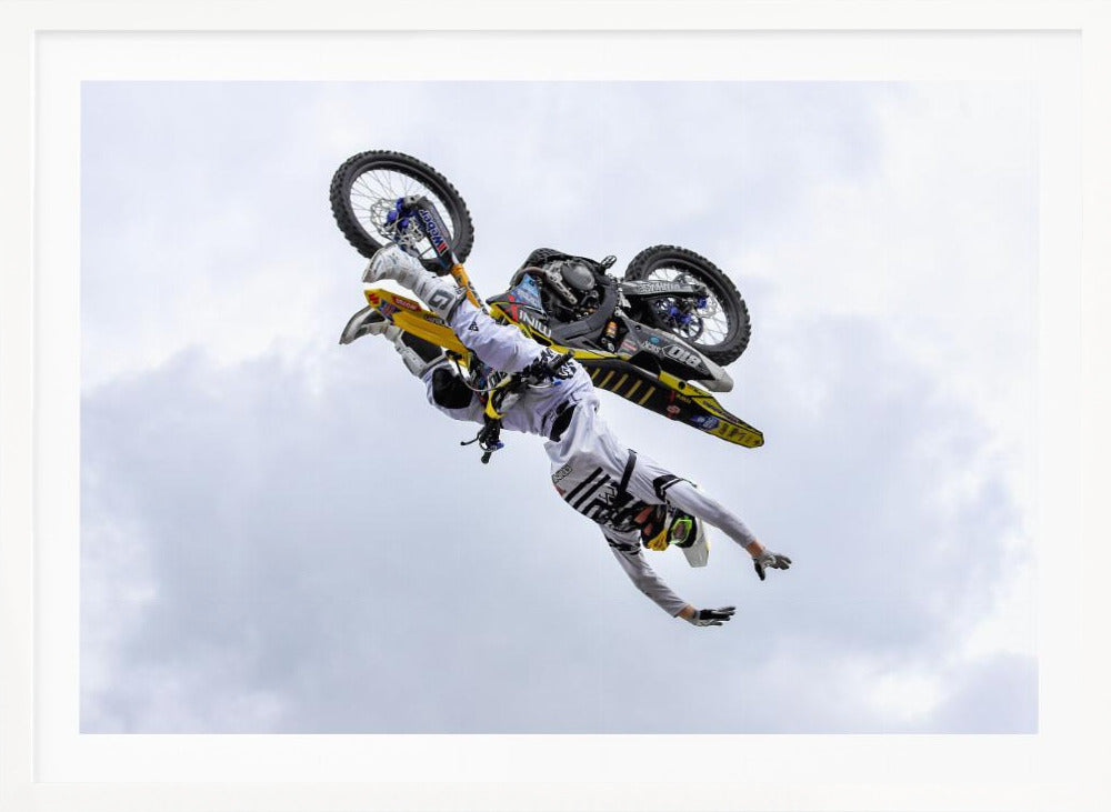 A framed action shot of a freestyle motocross rider in mid-air, performing an upside-down trick against a cloudy sky, with their body hanging below the yellow and black dirt bike. Artwork