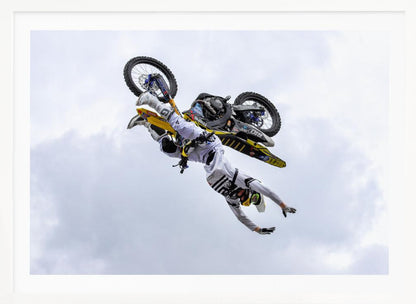 A framed action shot of a freestyle motocross rider in mid-air, performing an upside-down trick against a cloudy sky, with their body hanging below the yellow and black dirt bike. Artwork