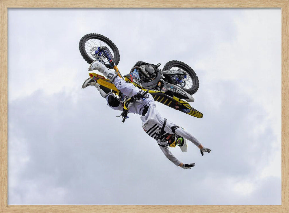 A framed action shot of a freestyle motocross rider in mid-air, performing an upside-down trick against a cloudy sky, with their body hanging below the yellow and black dirt bike. Artwork