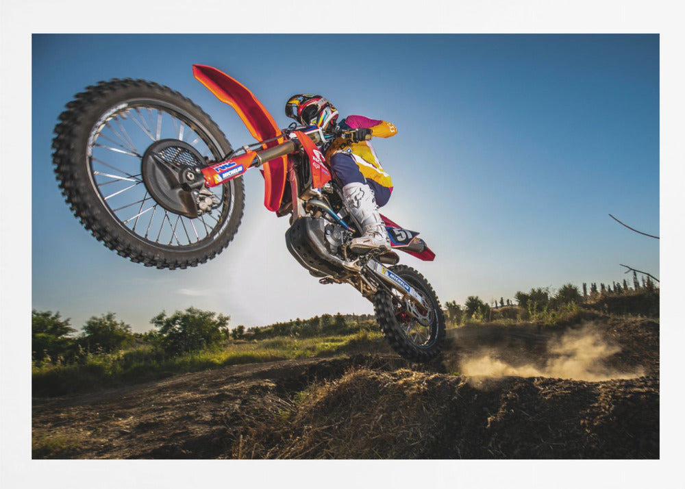 A dynamic, low-angle shot of a motocross rider in mid-air, jumping over a dirt mound. The rider is on a red, white, and blue dirt bike, wearing colorful racing gear and a helmet. The bike's front wheel is high in the air against a clear blue sky, while the back wheel is just clearing the jump, kicking up a cloud of dust. The background features a green, rural landscape. Wall Art