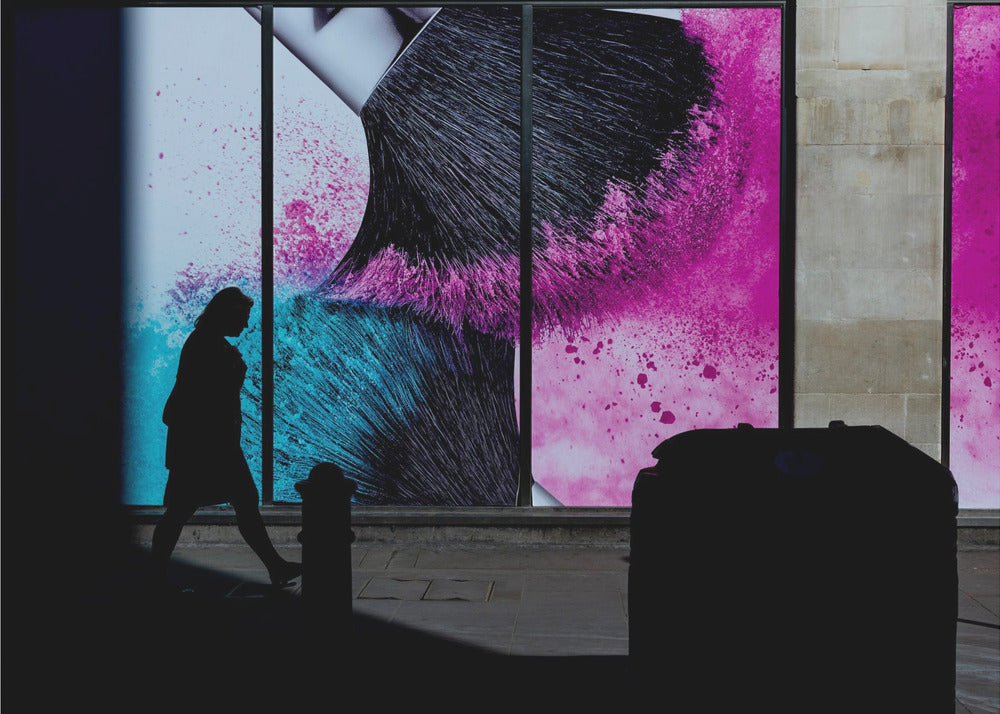 A framed photograph of a street scene where a person's silhouette is captured walking past a large, vibrant window advertisement. The ad features a close-up of a makeup brush with explosions of magenta and cyan powder. The foreground is cast in deep shadow, creating a dramatic contrast. Print