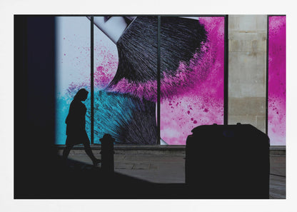 A framed photograph of a street scene where a person's silhouette is captured walking past a large, vibrant window advertisement. The ad features a close-up of a makeup brush with explosions of magenta and cyan powder. The foreground is cast in deep shadow, creating a dramatic contrast. Print