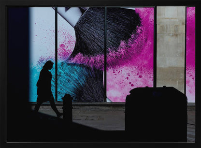 A framed photograph of a street scene where a person's silhouette is captured walking past a large, vibrant window advertisement. The ad features a close-up of a makeup brush with explosions of magenta and cyan powder. The foreground is cast in deep shadow, creating a dramatic contrast. Print