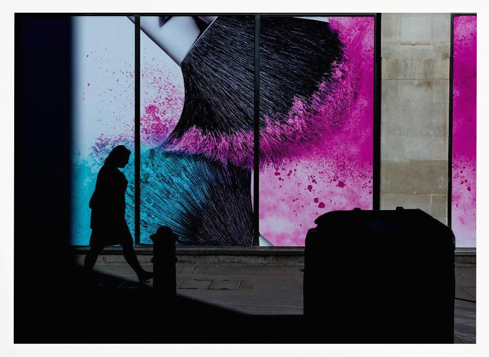 A framed photograph of a street scene where a person's silhouette is captured walking past a large, vibrant window advertisement. The ad features a close-up of a makeup brush with explosions of magenta and cyan powder. The foreground is cast in deep shadow, creating a dramatic contrast. Print