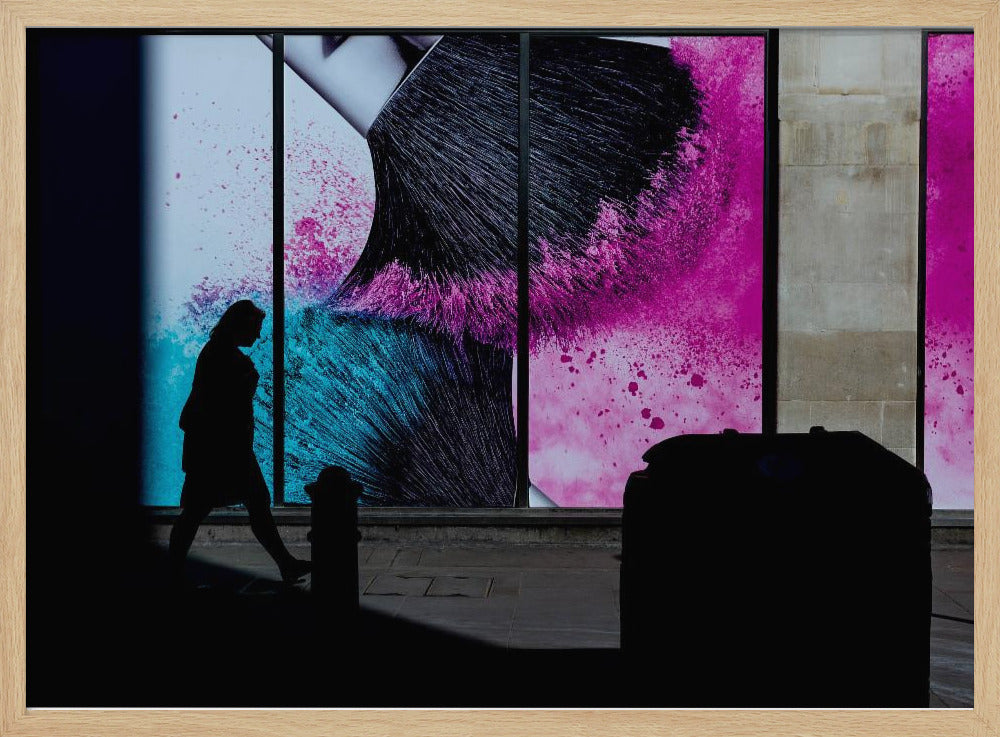 A framed photograph of a street scene where a person's silhouette is captured walking past a large, vibrant window advertisement. The ad features a close-up of a makeup brush with explosions of magenta and cyan powder. The foreground is cast in deep shadow, creating a dramatic contrast. Print
