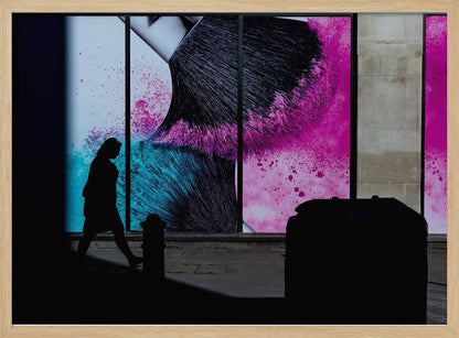 A framed photograph of a street scene where a person's silhouette is captured walking past a large, vibrant window advertisement. The ad features a close-up of a makeup brush with explosions of magenta and cyan powder. The foreground is cast in deep shadow, creating a dramatic contrast. Print