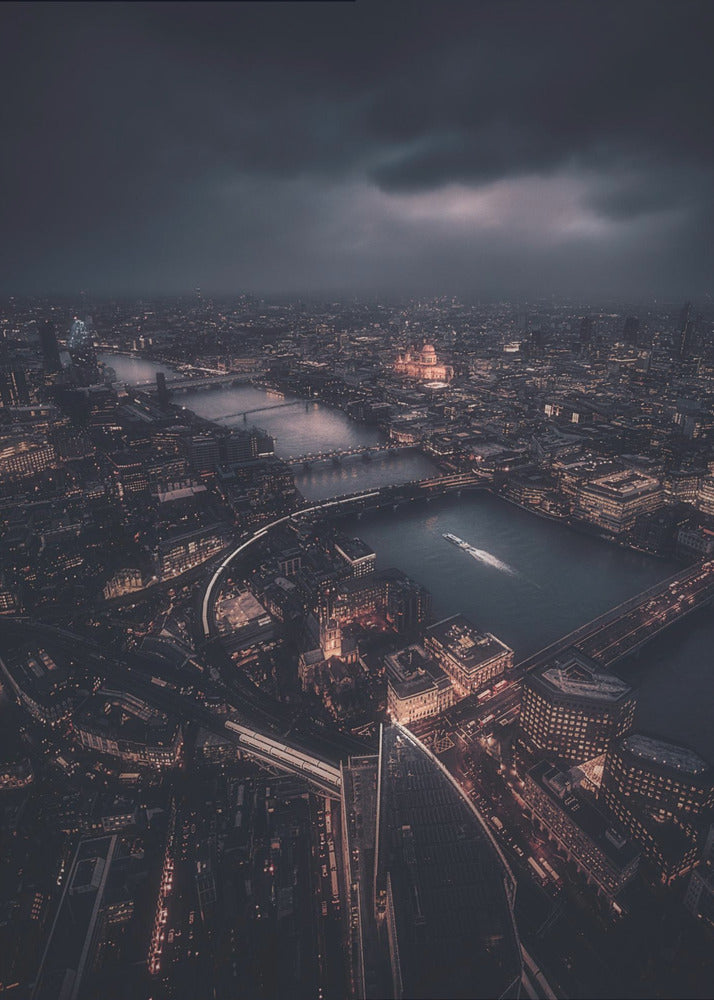 An aerial photograph of London at night from a high vantage point, overlooking the winding River Thames illuminated by the golden glow of city lights under a dark, dramatic, and cloudy sky. Wall Art