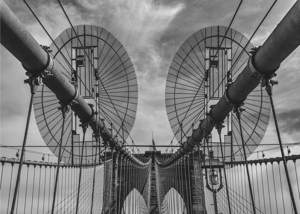 A symmetrical, low-angle black and white photograph of the Brooklyn Bridge's suspension cables and towers against a cloudy sky, framed in silver. Poster