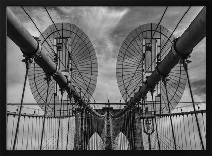 A symmetrical, low-angle black and white photograph of the Brooklyn Bridge's suspension cables and towers against a cloudy sky, framed in silver. Poster