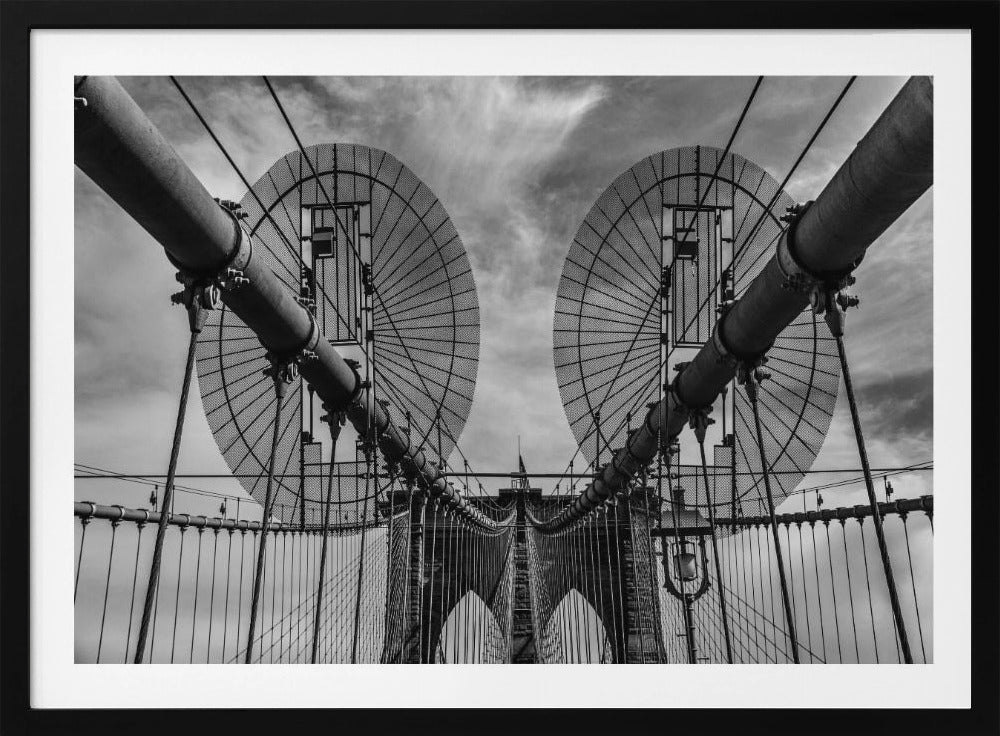 A symmetrical, low-angle black and white photograph of the Brooklyn Bridge's suspension cables and towers against a cloudy sky, framed in silver. Poster